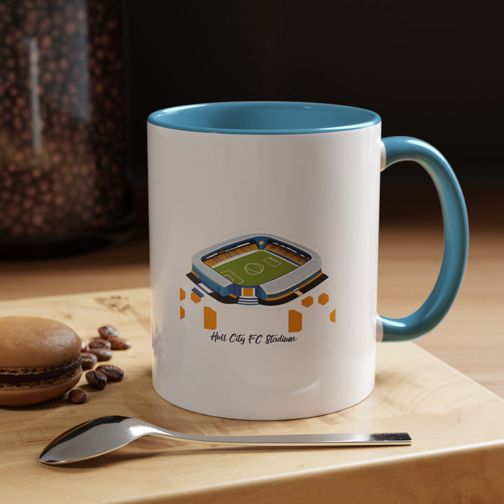 A blue Hull City FC stadium mug displaying a panoramic illustration of MKM Stadium printed with high clarity. The calming blue tone pairs well with the artwork, seen here in a relaxed workspace setting, capturing the comfort of enjoying tea or coffee while staying connected to the passion of supporting Hull City throughout the day.