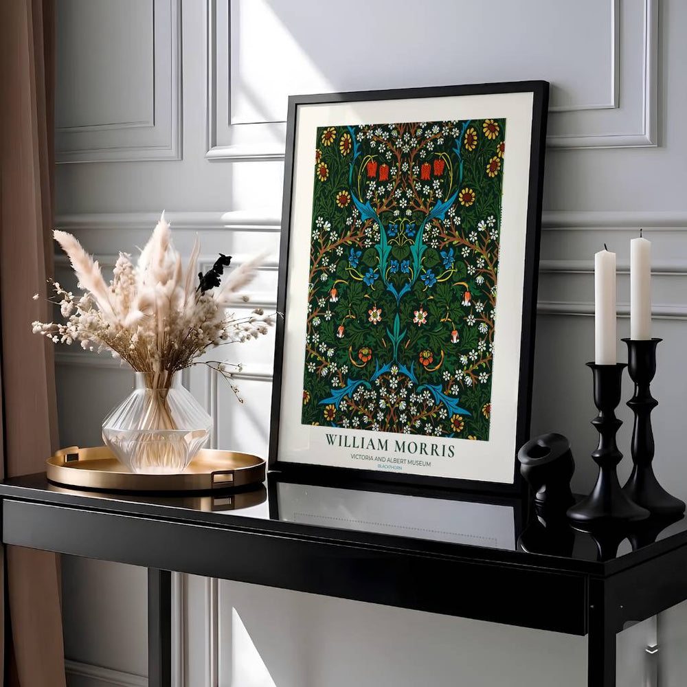 A framed William Morris art print featuring a detailed green and blue floral tree design, styled on a black console table next to a vase of dried grass and black candlesticks against a gray paneled wall.