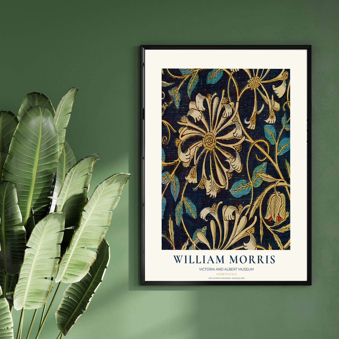 A framed William Morris art print featuring a navy blue background with cream, gold, and teal floral motifs, hung on a dark green wall next to large green leaves.
