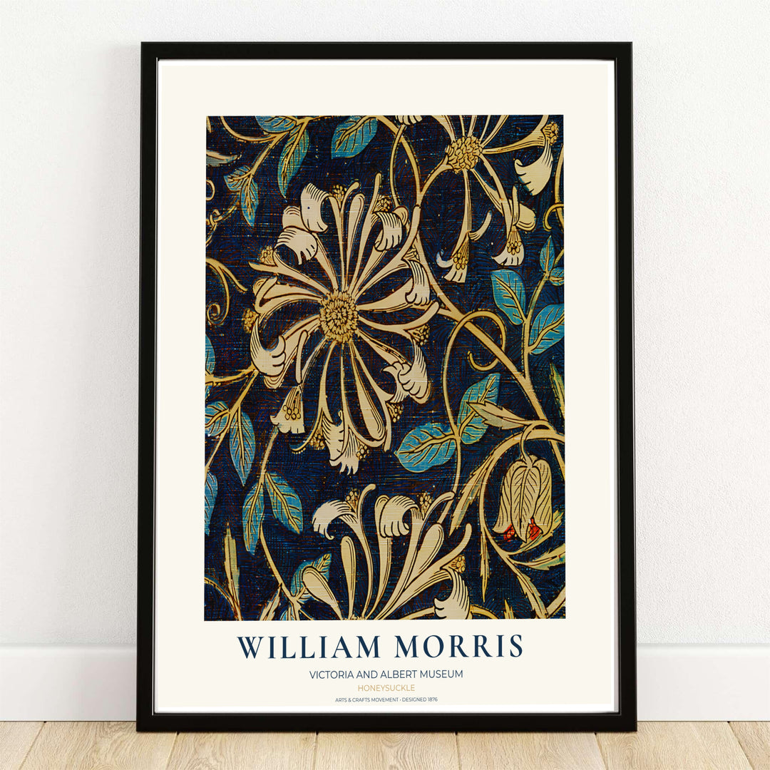 A framed William Morris artwork leaning against a white wall on a light wood floor. The print features an ornate floral pattern in cream, gold, and teal on a dark navy background, with the text &