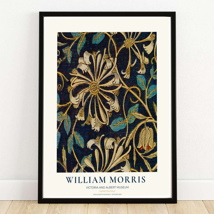 A framed William Morris artwork leaning against a white wall on a light wood floor. The print features an ornate floral pattern in cream, gold, and teal on a dark navy background, with the text &