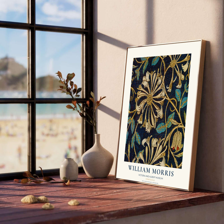 A framed William Morris art print with a floral pattern leans against a wall on a wooden windowsill. The print features cream and gold floral motifs on a dark navy background.