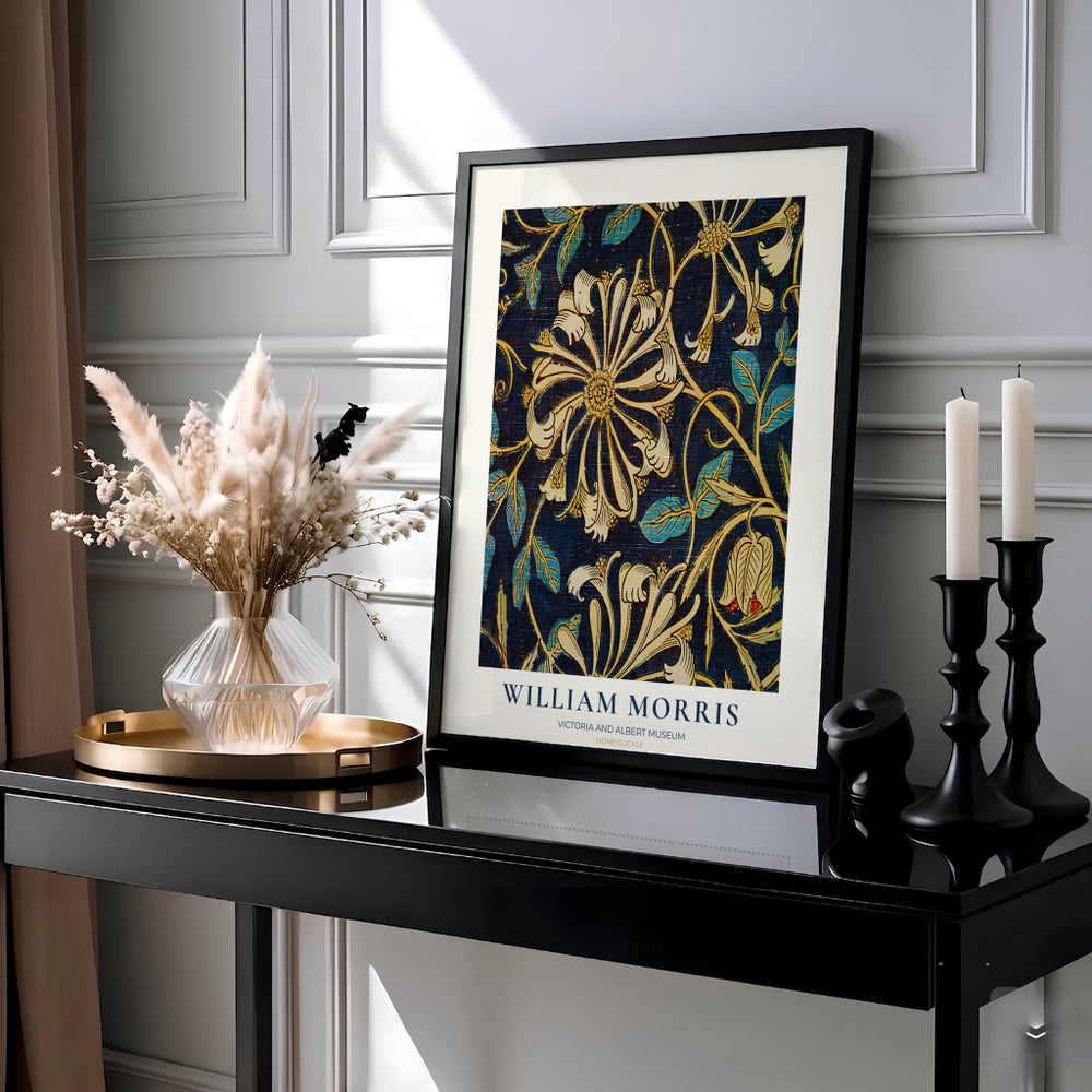 A framed William Morris botanical art print with gold and teal floral motifs on a dark navy background, styled on a black console table with dried flowers, candles, and decorative objects against a paneled gray wall.