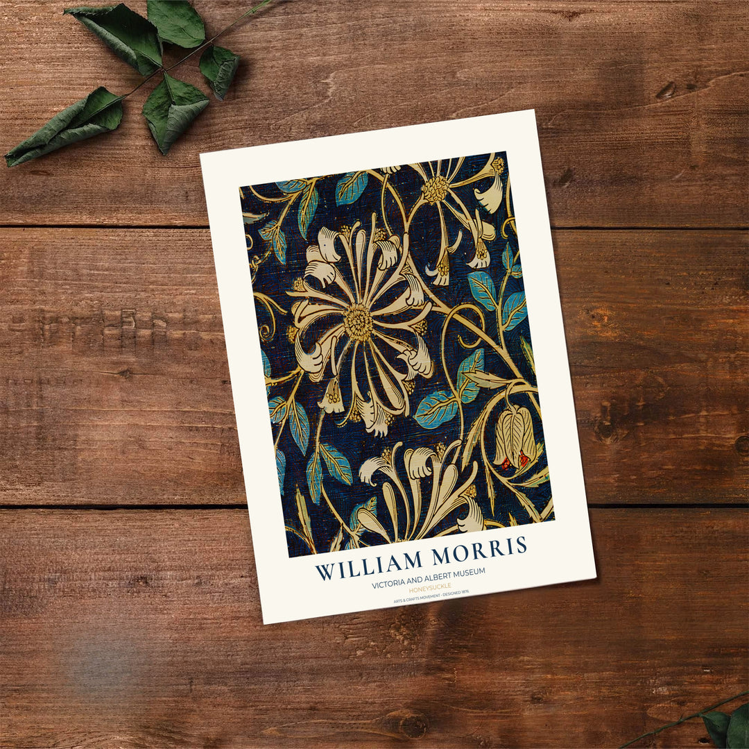 A William Morris art print featuring a floral pattern in cream, gold, and teal on a dark navy background, styled on a rustic wooden table with green leaves in the corner.