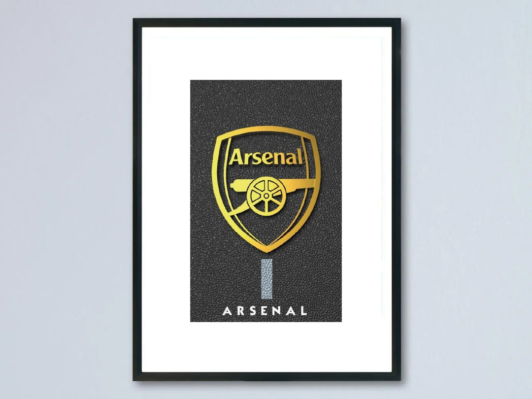 Framed metallic gold Arsenal FC crest poster featuring a shiny golden club shield and cannon logo over a premium textured pebble black background, showcased inside a modern black photo frame.