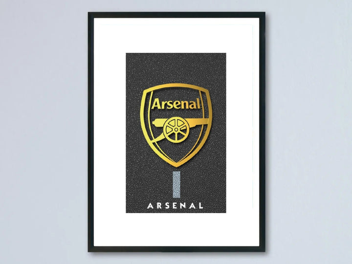 Framed metallic gold Arsenal FC crest poster featuring a shiny golden club shield and cannon logo over a premium textured pebble black background, showcased inside a modern black photo frame.