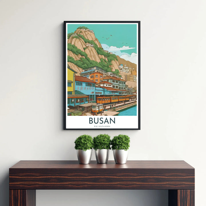 Picturesque Korean coastal art print highlighting a yellow and orange beach train passing in front of a hillside village in Busan, South Korea, under a clear teal sky. Offered by MapYourDreams, this artwork is available in flexible options including unframed paper, custom wooden frames, and gallery canvas wraps.